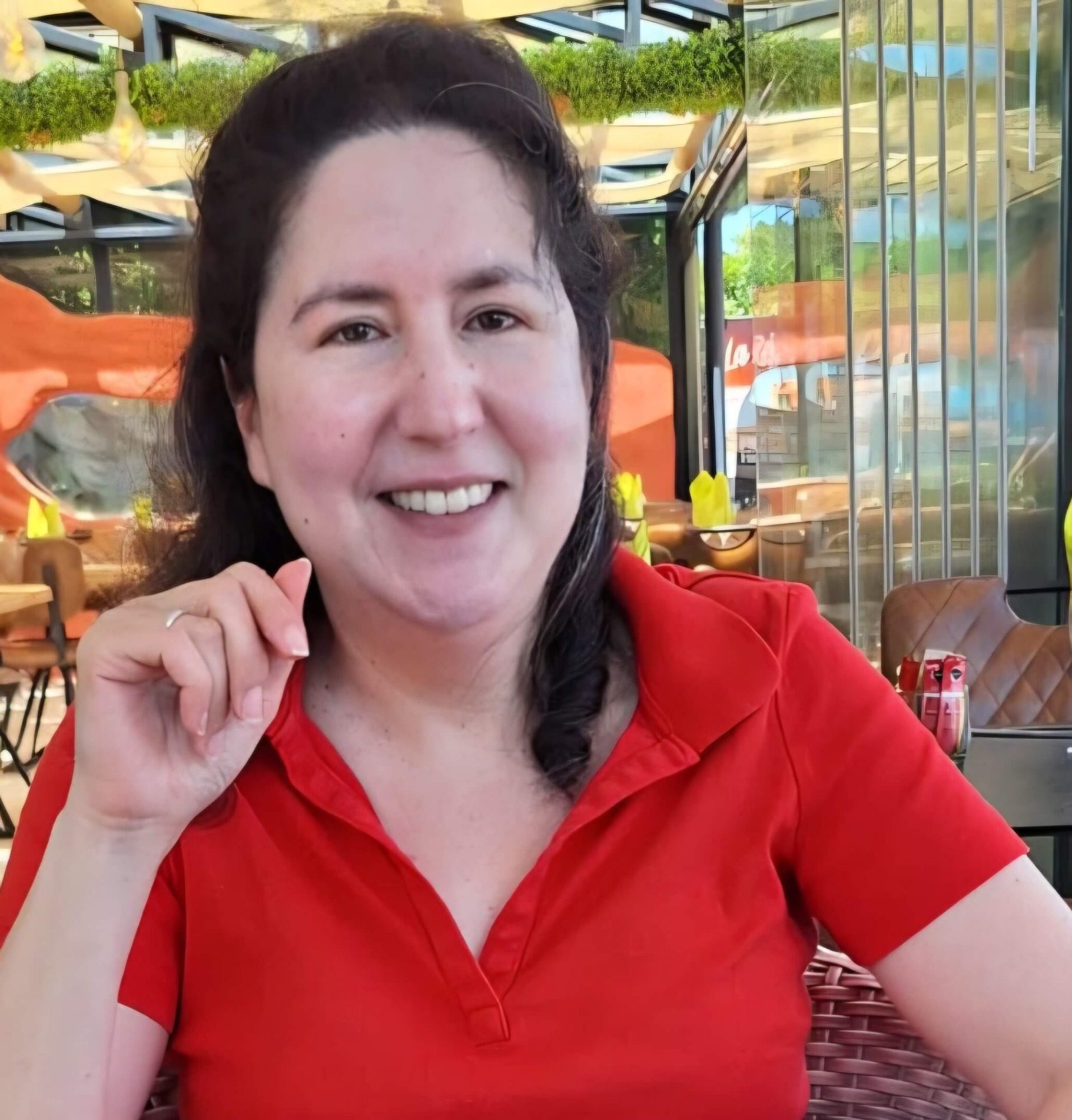 Vrouw in een rode blouse zit op een zonnig terras en lacht vriendelijk.